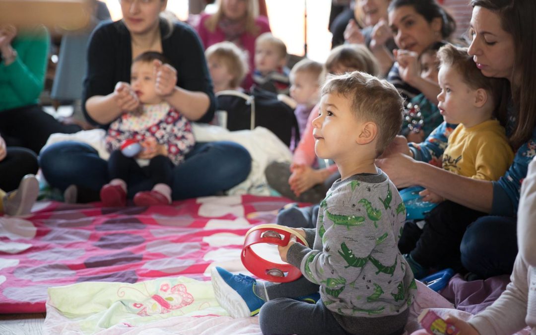 image shows little boy with tambourine in amongst a group of seated parents with babies