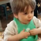 a little baby boy is sitting in his high chair with a green bib on; he is having a sign language conversation about how much he loves his daddy and his dog and showing his baby signing for love - tiny hands on his heart) to his mummy (not visible)