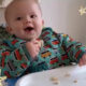a little baby boy is sitting in his high char having something to eat. He is wearing a green babygro with little red trains on it. He is smiling and happy, with food in his hand and on his face. He is showing his mummy (not visible) how he baby signs more