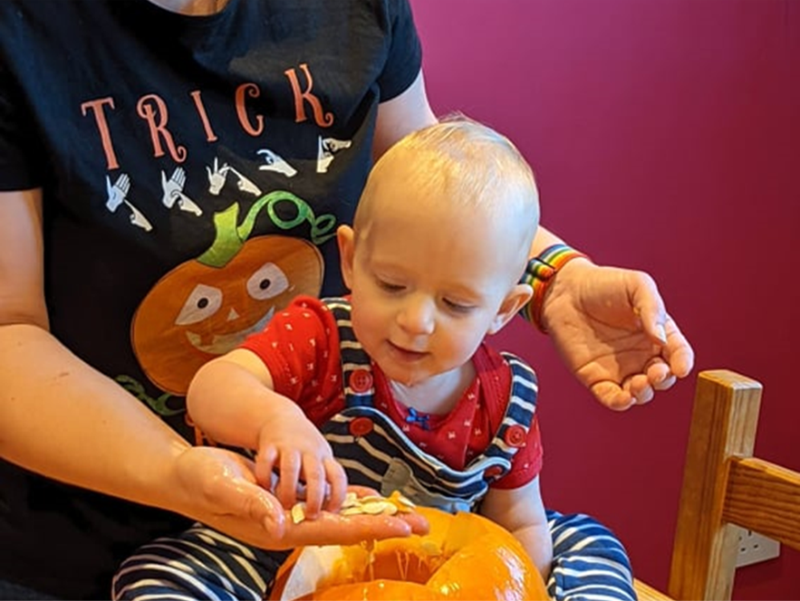 a blonde caucasian baby girl dressed in a red t-short and blue stripey dungarees is exploring an orange pumpkin with her mummy.  Her mummy is holding the seeds in her hand for her baby to touch and explore.  Mummy is wearing a sign language Trick or Treat t-shirt, which is black with the words Trick or Treat spelled out in British Sign Language fingerspelling and a fun motif of halloween pumpkin.